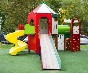 A wooden slide of bright red, blue and green color against a background of green trees and residential buildings on a clear sunny day. Playgrounds, sports, health entertainment.