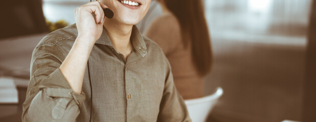 Young dark-haired guy in a green shirt and headsets is talking to a client, while sitting at the desk, working together with a female colleague in a modern office. Call center operators at work