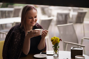 Close-up portrait of smiling woman drinking coffee alone. Lovable lady standing near window and looking at morning empty street with cup of coffee.