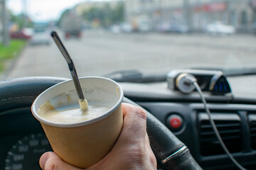 close up, the driver hand holds a disposable cardboard cup with foam coffee, cocoa and a straw inside the car against the background of the roadway, the highway in the evening