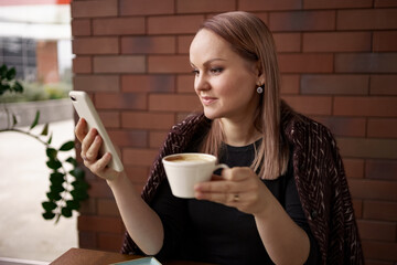 Close-up portrait of smiling woman drinking coffee alone while talking on smartphone. Lovable lady standing near brick wall in a cafe