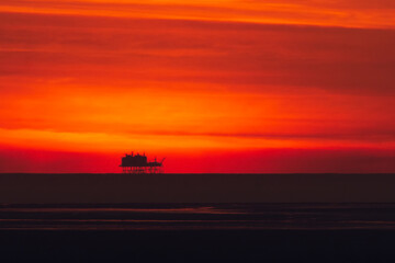 Fototapeta na wymiar Distant oil rig or platform silhouette against orange sky at sunset. Theme of petrochemicals, sunset industry, industry, business