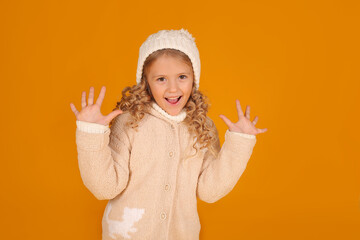 a beautiful little surprised blonde girl in a knitted jacket and a winter knitted hat on a yellow...