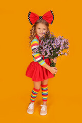 a little blonde girl with a red bow on her head in a red skirt and a colored blouse and knee socks on a yellow background with flowers in her hands