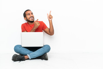 Young Ecuadorian man with a laptop sitting on the floor isolated on white background pointing with...