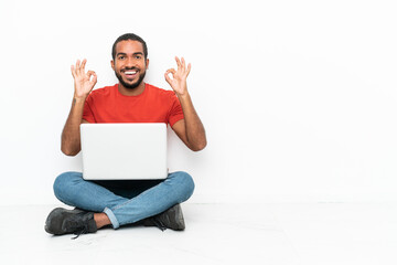 Young Ecuadorian man with a laptop sitting on the floor isolated on white background showing an ok sign with fingers