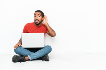 Young Ecuadorian man with a laptop sitting on the floor isolated on white background listening to something by putting hand on the ear