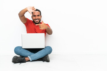 Young Ecuadorian man with a laptop sitting on the floor isolated on white background focusing face. Framing symbol