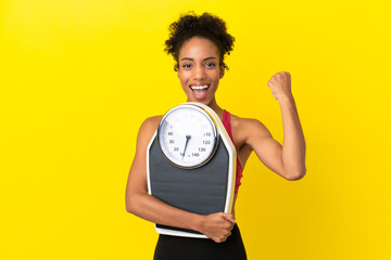 Young African American woman isolated on yellow background with weighing machine and doing victory gesture
