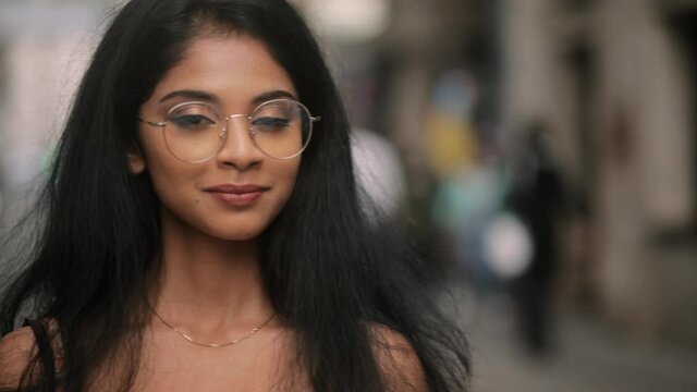 Portrait of attractive Indian girl in eyeglasses looking at the camera. Young girl with long hair at the city street. . High quality 4k footage