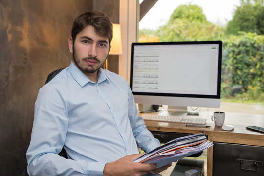 jeune homme au travail ou en t&eacute;l&eacute;travail devant son ordinateur tenant dans ses mains une pile de dossiers
