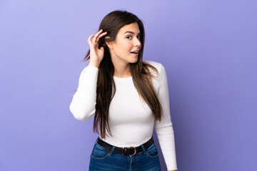Teenager Brazilian girl over isolated purple background listening to something by putting hand on the ear