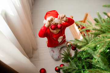 Fototapeta premium little boy in red pajamas decorates the Christmas tree with toys and balls. Cute kid preparing home for xmas celebration. winter New Year's concept. space for the text. High quality photo