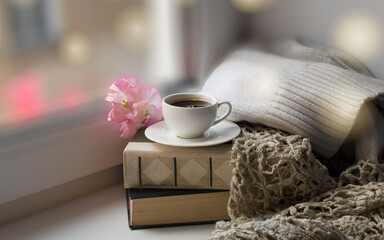 A white cup with coffee on a saucer on a stack of books, a knitted shawl, pink sweet pea flowers, a...