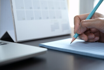 Close-up of a business woman hand writing notes on paper notepad, making agenda on personal organizer with desktop calendar and laptop computer on office table at workplace