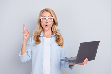 Portrait of shocked amazed mature businesswoman advertise product hold laptop wear glasses isolated on grey color background