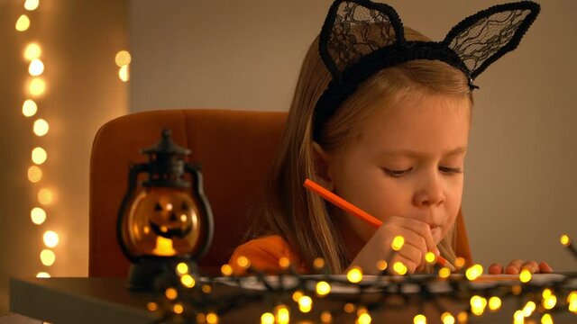 Close up of little girl sits in dark and makes decoration for holiday halloween. Child with pumpkin lamp on background of shining blurred lights of golden garland.