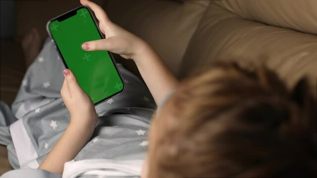 little girl schoolgirl lies relaxed on the couch and uses a smartphone with a green screen chroma key, scrolls up