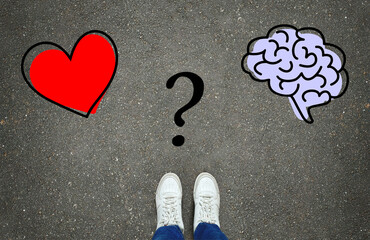Heart or brain? Legs in white sneakers in front of heart and brain symbols with a question mark between them on the gray asphalt background. Path choice concept