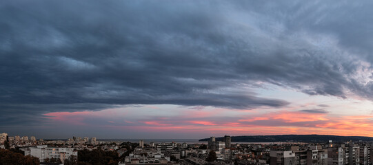 Stormy gray clouds is coming to seaside city at sunset. Panorama of dramatic overcast sky at evening. Dark stratus clouds hang over Varna. Weather change concept. Storm is coming wide landscape.  - obrazy, fototapety, plakaty