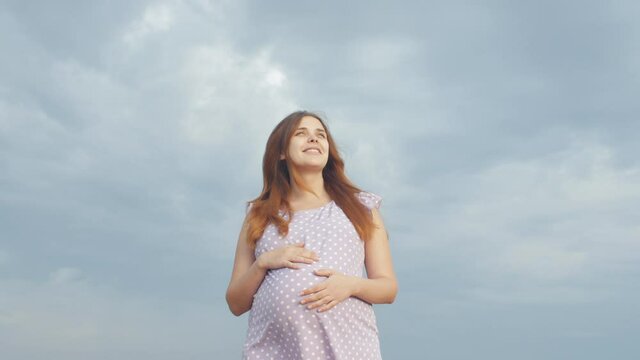 camera rotates around happy pregnant woman in dress standing alone in field on background of cloudy rainy sky