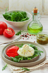 Creamy Italian Burrata Cheese Served with fresh arugula, Tomatoes, Olive Oil, Basil Pesto and freshly ground pepper on the grey Plate on a white wooden background. Selective focus