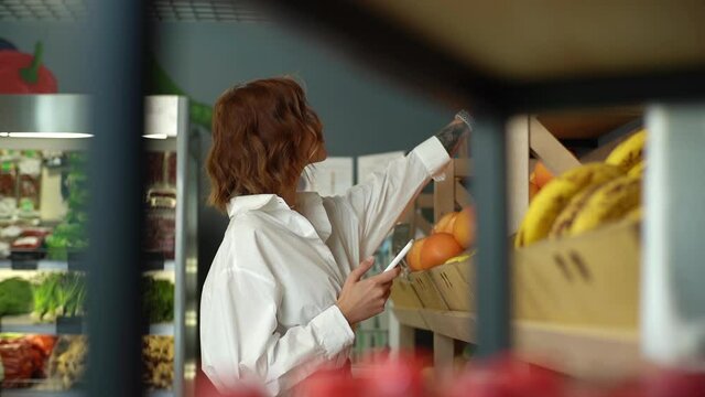 Female choosing products in market walking along counters taking photos of items using smartphone, slow motion. Side view of young woman at grocery store looking up nutrition facts using mobile phone.