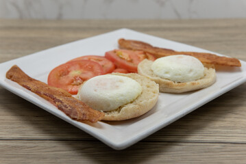 Poached eggs sitting on english muffin served with bacon and sliced tomato for a hearty breakfast