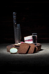 dark background. round wooden bathtub in retro style. a piece of bacon is cut into pieces. a sharp knife made of forged steel. black bread, a head of onion and a glass of vodka.