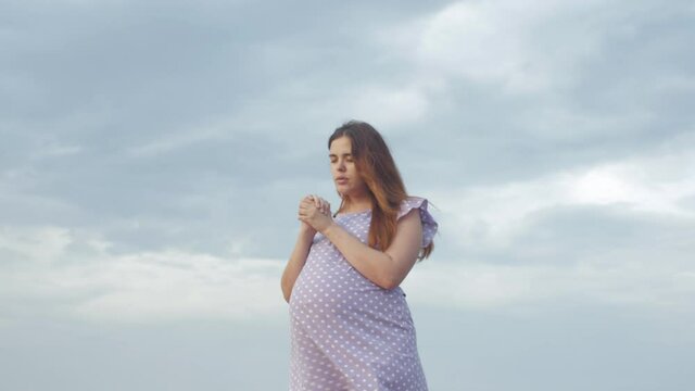 young pregnant woman folded her hands in pray to God standing alone in field against of cloudy sky, future mother walking on nature