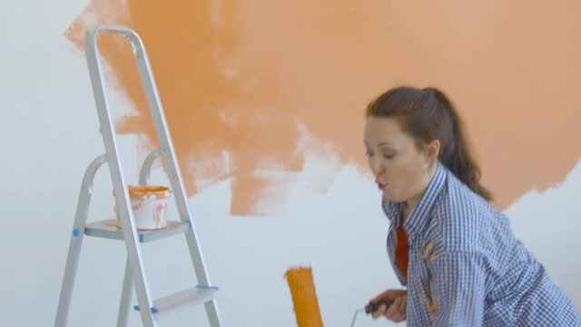 Singleton lifestyle and renovation concept. Young smiling woman with painting roller paints the wall in the apartment and dances