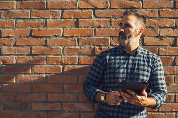 Portrait of modern businessman  with beard thinking and  using digital tablet while  standing in front of brick wall  outdoor.