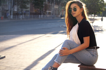 Young pretty brunette woman wearing casual clothes and sunglasses on the background of the city in the rays of the setting sun.