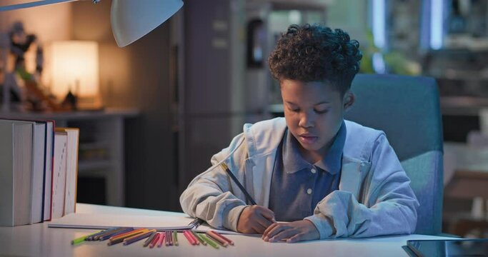 Cheerful African american boy artist playing alone drawing coloring picture with pencils at night, focused smart preschool child enjoy creative art hobby activity at home, children development concept