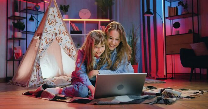 Attractive happy smiling carefree two diverse girls sitting on the floor on coverlets near wigwam and use laptop to watch interesting program in the evening at home