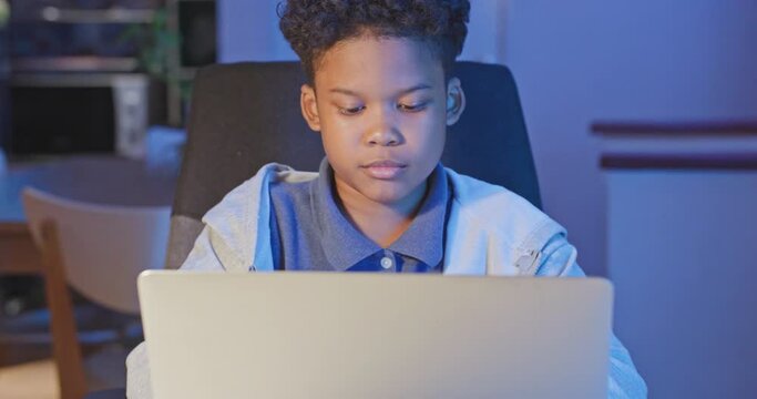 African american boy use computer laptop to studying online at night or doing schoolwork in living room at home while city lockdown. Social distancing during covid 19 and New normal concept.