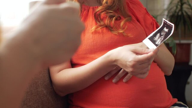 pregnant young woman sitting in kitchen with girlfriend talking about ultrasound snapshot, motherhood concept, pregnancy and friendship
