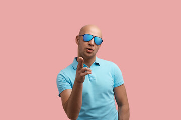 Playful handsome bald unshaven homosexual man makes shooting gesture towards camera, shoots target, keeps mouth open, gay friendly, wears blue polo shirt and sunglasses, stands over pink background.