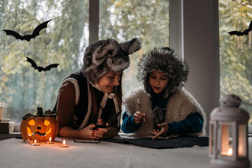 Mom and son in Halloween costumes play together and make paper bat Halloween decorations on pumpkins lamp at home backgound. Halloween family at home, Halloween parties and decorations