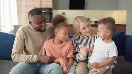 Portrait of beautiful diverse family of Black man, White woman and two cute biracial children talking cheerfully sitting on sofa at home. Multiethnic family enjoying spending time together