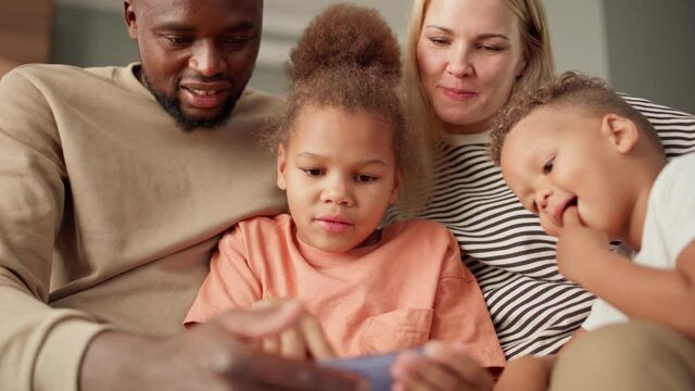 Cute multiracial family of four using smartphone together at home. Biracial little girl explaining diverse parents and toddler brother how to play mobile video game on cell phone