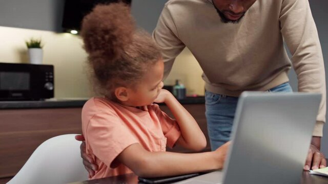 Biracial elementary student doing homework or learning online at home struggling to fulfill task. Caring Black father helping girl with study, explaining lesson to daughter. Open laptop on table