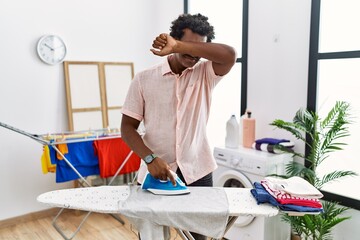 African man with curly hair ironing clothes at home smiling cheerful playing peek a boo with hands...