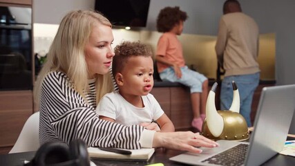 Busy Caucasian mother working on laptop from home and talking to biracial toddler son sitting on her lap and interrupting. Black father cooking and talking with daughter behind. Family staying at home - Powered by Adobe