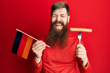 Redhead man with long beard holding fork with pork sausage and germany flag smiling and laughing...