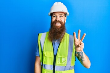 Redhead man with long beard wearing safety helmet and reflective jacket showing and pointing up with fingers number three while smiling confident and happy.