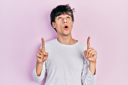 Handsome hipster young man wearing casual white shirt amazed and surprised looking up and pointing with fingers and raised arms.