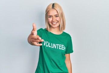 Beautiful blonde woman wearing volunteer t shirt smiling friendly offering handshake as greeting and welcoming. successful business.