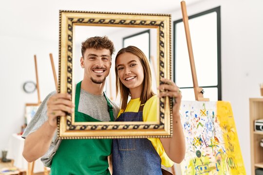 Young hispanic artist couple smiling happy holding empty frame at art studio.