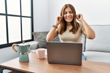 Young brunette woman using laptop at home drinking a cup of coffee covering ears with fingers with annoyed expression for the noise of loud music. deaf concept.